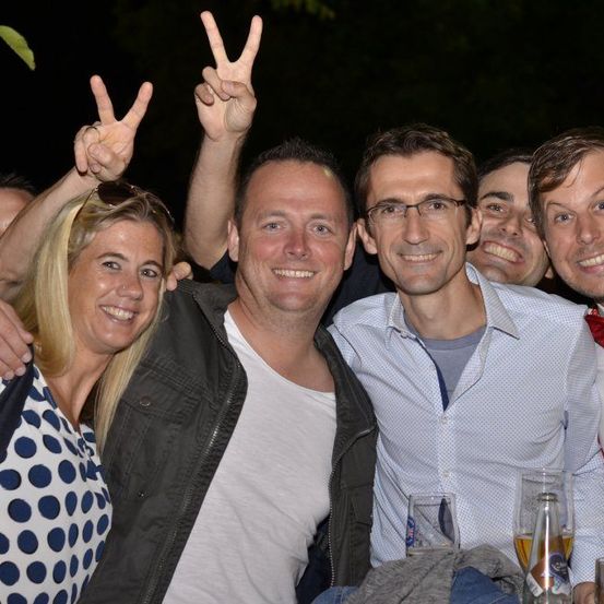 Eine Gruppe von Menschen, die nachts lächelnd für ein Foto posieren, einer mit Brille und einer mit Fliege, mit Getränken vor ihnen.