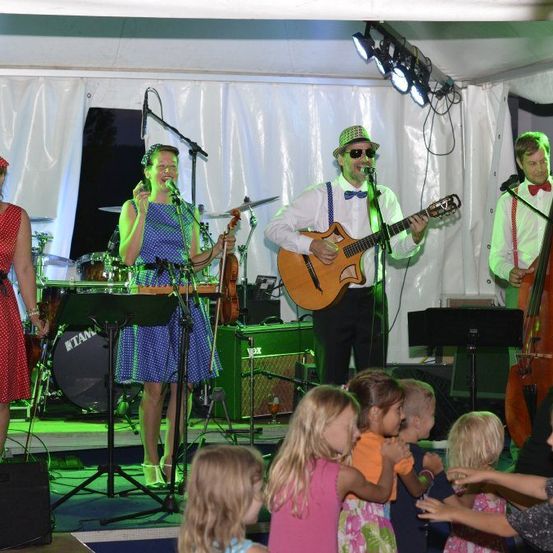 Eine Band tritt auf einer Bühne mit grüner Beleuchtung auf. Die Band besteht aus einem Geiger, einem Gitarristen, einem Cellisten und zwei Sängern. Kinder tanzen vor der Bühne.