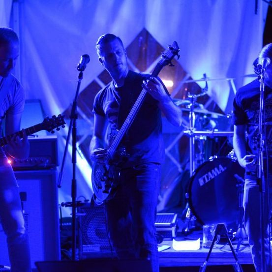 Eine Band spielt auf der Bühne bei blauer Beleuchtung. Der Gitarrist links spielt, während der Schlagzeuger hinter einem Tama-Schlagzeug steht. Der Gitarrist in der Mitte spielt Bassgitarre.