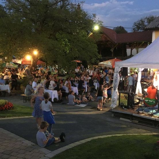 Eine große Menschenmenge versammelt sich für eine Veranstaltung im Freien, mit Menschen, die an Tischen unter Schirmen sitzen, und anderen, die eine Bühnenaufführung beobachten. Die Atmosphäre ist lebhaft mit tanzenden Kindern und sozialisierenden Erwachsenen. Bäume und ein Gebäude bieten einen malerischen Hintergrund.