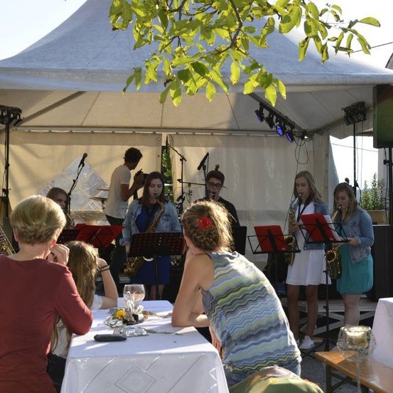 Eine Gruppe von Musikern spielt auf der Bühne unter einem weißen Zelt, während die Besucher an Tischen davor sitzen. Die Musiker spielen Saxophone und das Publikum hört aufmerksam zu.