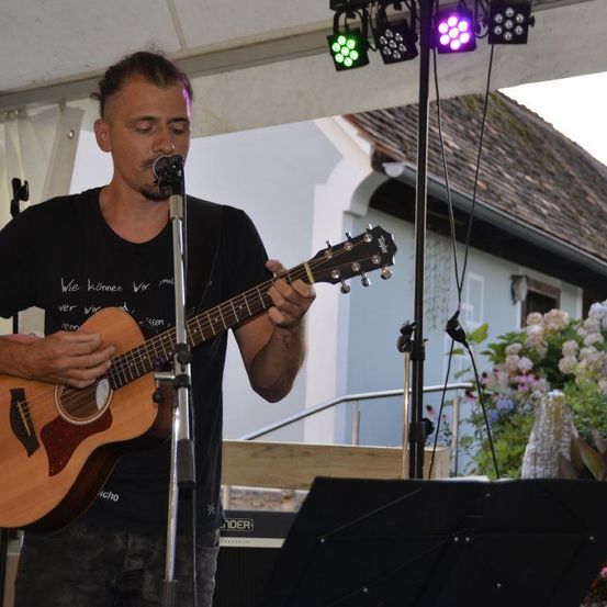 Ein Mann spielt Gitarre und singt in ein Mikrofon auf einer Bühne. Die Bühne hat ein weißes Zelt, bunte Lichter und im Hintergrund ein Haus mit Blumen.