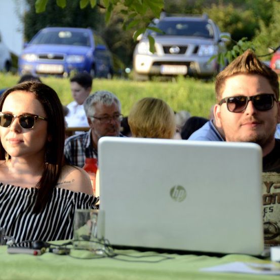 Ein Mann und eine Frau sitzen mit einem offenen Laptop an einem Tisch. Sie tragen Sonnenbrillen. Dahinter sitzen mehrere Personen auf einer Rasenfläche. Im Hintergrund sind Autos geparkt.