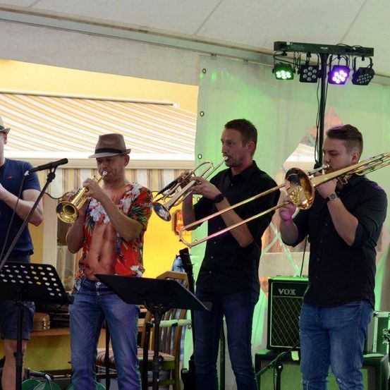 Vier Männer spielen Trompeten und Posaunen auf einer Bühne, einer trägt einen Hut und ein Blumenhemd, unter grüner Beleuchtung.