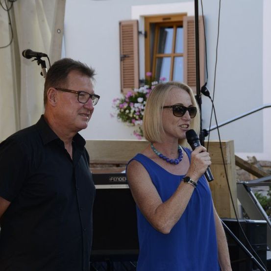 Ein erwachsener Mann und eine Frau stehen neben einem Mikrofon. Der Mann trägt ein schwarzes Hemd, eine Brille und ein Lächeln. Die Frau trägt ein blaues ärmelloses Top, eine Sonnenbrille und eine Halskette. Sie stehen wahrscheinlich auf der Bühne, mit einem Gebäude, einer Treppe und einem Mikrofonständer dahinter.