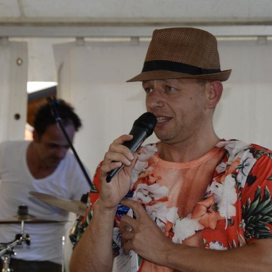 Ein Mann in einem Hut singt in ein Mikrofon, während ein anderer Mann Schlagzeug spielt.