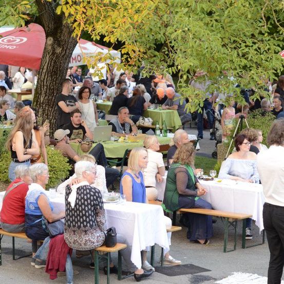 Viele Menschen sitzen im Freien in einem Park an Tischen. Einige sitzen auf Bänken, andere auf Stühlen. Die Tische sind mit grünen Tischdecken bedeckt. Hinter den Tischen stehen Zelte. Es gibt Bäume in der Umgebung.