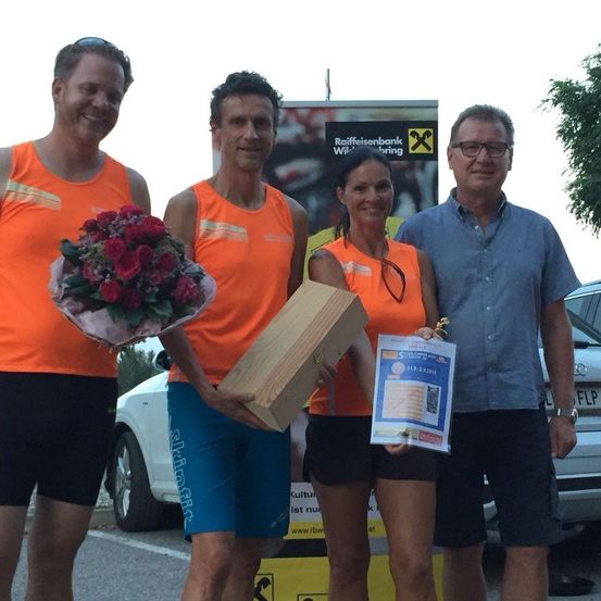 Vier Menschen stehen auf einem Parkplatz mit einem Gebäude und einem Banner dahinter. Die Frau hält eine Schachtel und ein Papier mit einem Logo. Der Mann auf der linken Seite hält einen Blumenstrauß. Der Mann auf der rechten Seite trägt eine Uhr. Dahinter ist ein Auto geparkt.