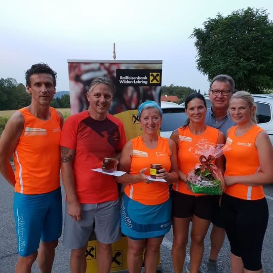 Fünf Personen in orangefarbenen Westen stehen vor einem Banner und posieren für ein Foto. Drei Frauen halten Geschenke, während zwei Männer kleine Gegenstände halten. Im Hintergrund befinden sich ein Banner, Bäume und ein geparktes Auto.