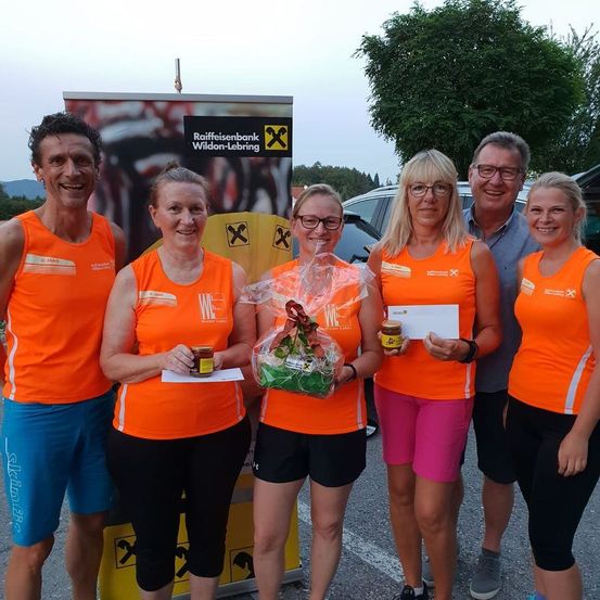 Fünf Personen in orangefarbenen Westen und Sportbekleidung posieren für ein Foto, halten Auszeichnungen und Blumen in der Hand, vor einem Banner mit dem Text 'Raiffeisenbank Wildon-Leibring'. Hinter ihnen sind ein Baum, ein Auto und ein Gebäude zu sehen.