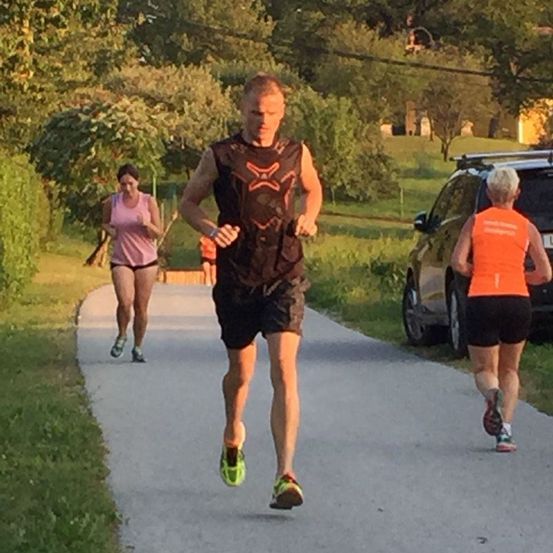 Drei Personen joggen auf einem gepflasterten Weg, umgeben von Grün und Bäumen. Ein Mann vorne trägt ein schwarzes Weste mit einem Sternmuster. Hinter ihm joggt eine Frau in einem rosa Tanktop. Eine weitere Frau folgt in einem orangefarbenen Shirt und joggt neben einem geparkten Auto.