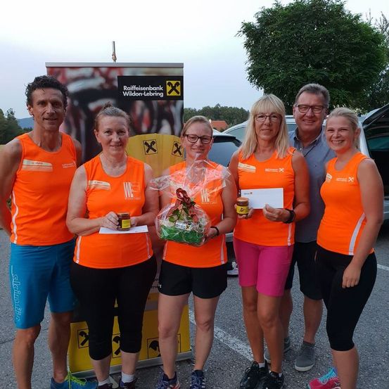 Fünf Personen in orangefarbenen Laufshirts stehen draußen. Zwei Frauen halten Blumen und Urkunden. Ein Mann in einem blauen Hemd und eine Frau in pinken Shorts halten ebenfalls Gegenstände. Ein Banner hinter ihnen lautet 'Raiffeisenbank Wildon-Leibring'.