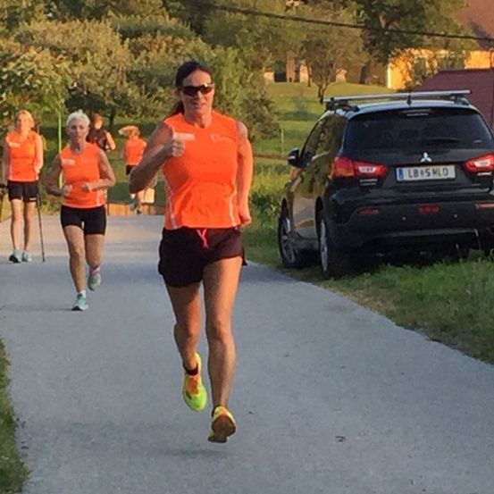 Drei Frauen laufen auf einem gepflasterten Weg, in der Nähe eines geparkten Autos. Sie tragen orangefarbene Shirts und schwarze Shorts. Das Auto hat ein Mitsubishi-Kennzeichen.