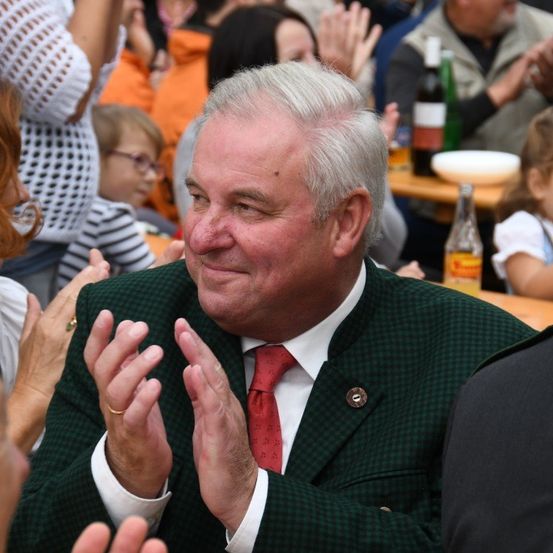 Ein älterer Mann in einem grünen Anzug und einer roten Krawatte klatscht bei einer Veranstaltung im Freien, umgeben von anderen Menschen, die ebenfalls klatschen.