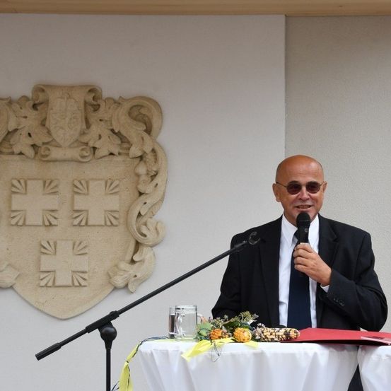 Ein Mann steht mit einem Mikrofon am Rednerpult, gekleidet in formeller Kleidung und lächelt. Hinter ihm befindet sich eine dekorative Wandplakette, und auf dem Tisch davor sind Blumen, Früchte und eine rote Mappe.