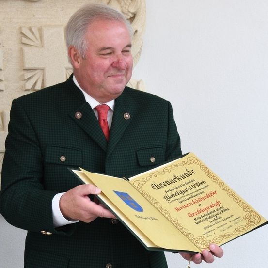 Ein Mann in einem grünen Anzug und roter Krawatte hält ein offenes Buch mit einem Zertifikat in der Hand und lächelt in die Kamera. Der Hintergrund zeigt eine Wand mit einem dekorativen Muster.