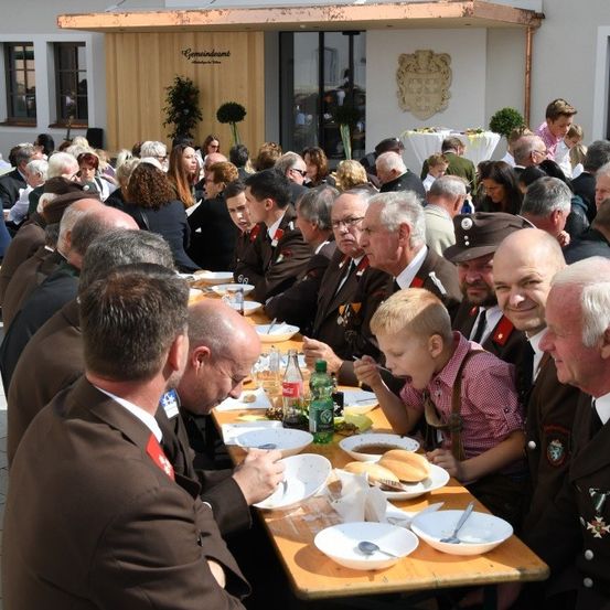 Eine große Gruppe von Menschen versammelt sich zu einer Mahlzeit bei einer Veranstaltung im Freien. Sie sitzen an langen Tischen, einige in Uniformen, und ein Kind isst mit einem Löffel.
