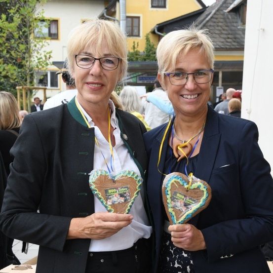 Zwei Frauen stehen nebeneinander im Freien und halten herzförmige Kekse. Sie tragen eine Brille und Jacken. Im Hintergrund ist ein Gebäude, Bäume und Menschen zu sehen.