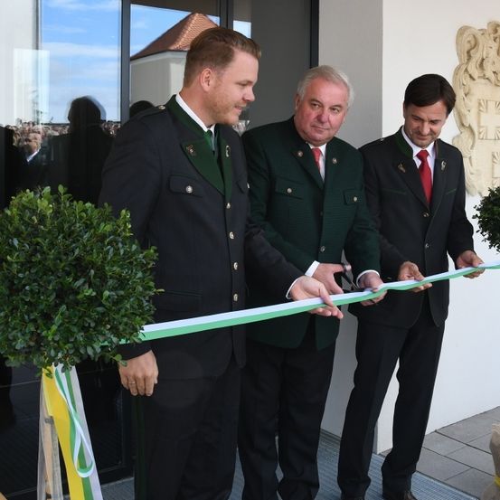 Drei Männer in Anzügen stehen vor einem Gebäude und schneiden ein Band, um es zu eröffnen. Zwei Männer tragen rote Krawatten.