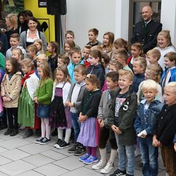 Eine Gruppe von Kindern und Erwachsenen steht auf einem gefliesten Boden, wahrscheinlich bei einer Veranstaltung. Einige Kinder tragen Uniformen und Schuhe, während andere Jacken und Sneaker tragen. Eine Frau und ein Mann stehen hinter ihnen. Ein Lautsprecher ist an der Wand montiert.