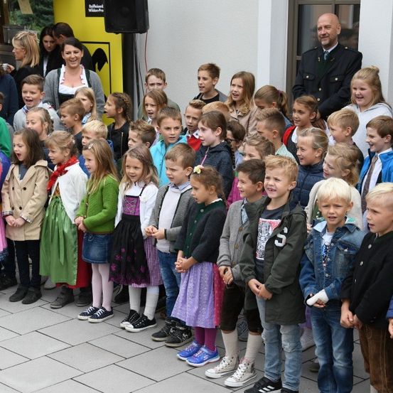 Eine Gruppe von Kindern und Erwachsenen steht auf einem gefliesten Boden, wahrscheinlich bei einer Veranstaltung. Einige Kinder tragen Uniformen und Schuhe, während andere Jacken und Sneaker tragen. Eine Frau und ein Mann stehen hinter ihnen. Ein Lautsprecher ist an der Wand montiert.