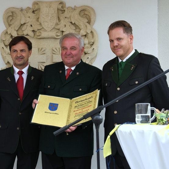 Drei Männer in Uniformen stehen vor einer Wand mit einem Wappen. Der Mann in der Mitte hält ein Zertifikat und der Mann rechts hat ein Glas Wasser auf einem Tisch.