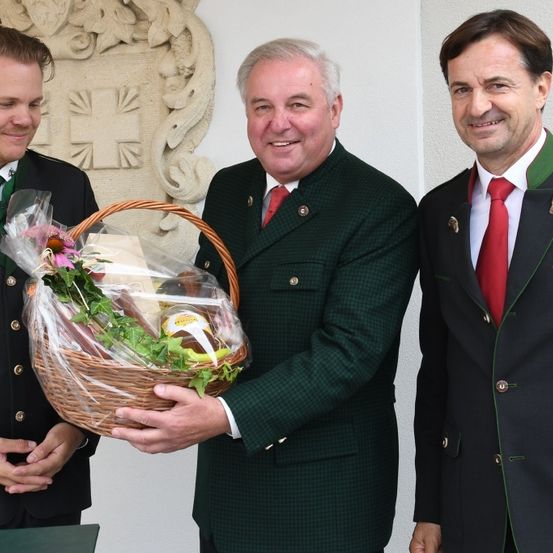 Drei Männer stehen nebeneinander und lächeln. Ein Mann hält einen Korb mit Blumen und Lebensmitteln, während die anderen beiden formelle Kleidung mit roten Krawatten und grünen Akzenten tragen. Sie scheinen für ein Foto zu posieren.