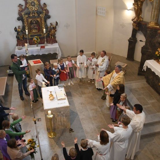 Eine religiöse Zeremonie findet in einer Kirche statt. Der Priester führt eine Gruppe von Kindern und Erwachsenen an. Altäre mit Kerzen und religiösen Symbolen schmücken die Wände.