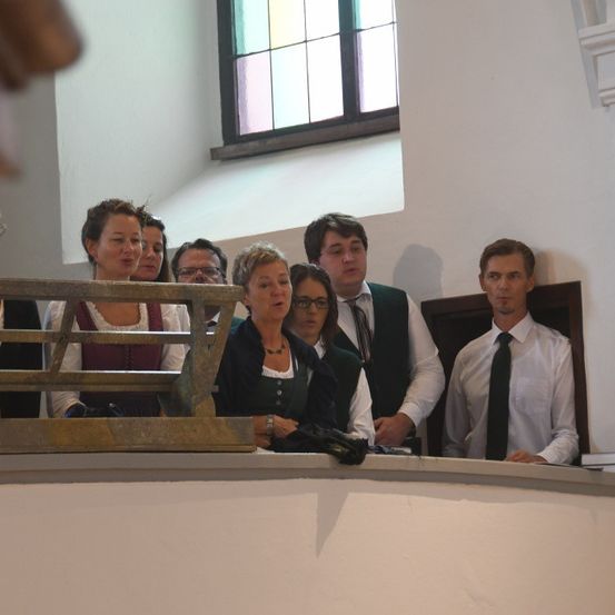 Mehrere Personen versammeln sich in einem Raum mit einer Holzbank, weißen Wänden und einem Buntglasfenster. Einige stehen, andere sitzen. Eine Frau hält eine Kamera, und ein Mann schaut sie an.