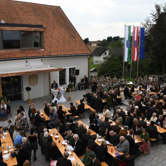Eine große Menschenmenge bei einer Gemeinschaftsveranstaltung, die an Tischen auf einer Terrasse sitzen, mit Fahnen und einem Gebäude im Hintergrund.