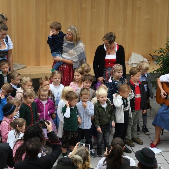 Eine Gruppe von Kindern und Erwachsenen versammelt sich für eine Veranstaltung im Freien. Eine Frau spielt Gitarre vor ihnen. Viele Kinder sind in traditioneller Kleidung gekleidet. Einige machen Fotos.