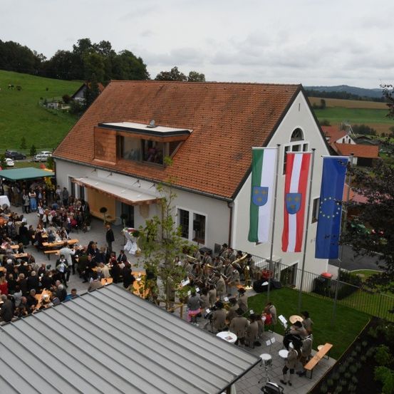Ein Luftbild eines Gebäudes mit braunem Dach und drei Fahnen. Mehrere Menschen sind draußen versammelt, möglicherweise für eine Zeremonie. Einige sitzen an Tischen und eine Brassband spielt auf dem Rasen.