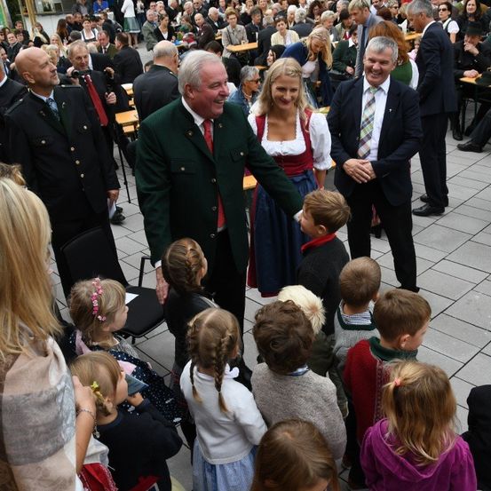 Eine Gruppe von Erwachsenen und Kindern steht und sitzt im Freien. Einige lächeln und reden, während andere zuschauen. Eine große Menschenmenge ist anwesend.