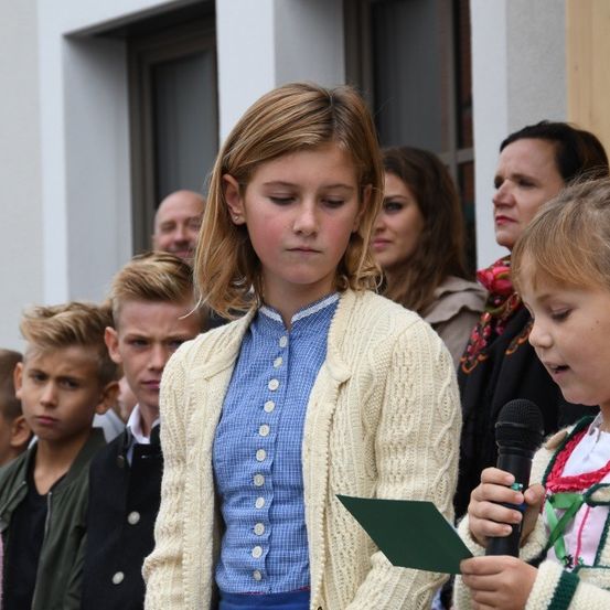 Ein junges Mädchen steht vor einer Menschenmenge und liest laut aus einem Stück Papier vor, während ein Mädchen neben ihr ein Mikrofon hält. Dahinter sind mehrere Erwachsene und Kinder versammelt, mit Gebäuden im Hintergrund.
