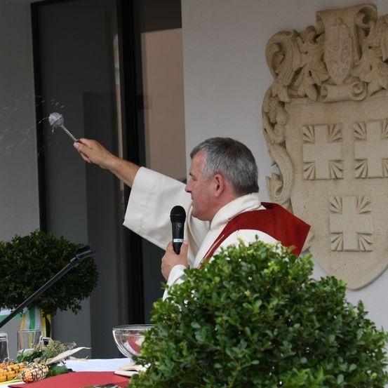Ein Priester sprüht Weihwasser aus einem silbernen Aspergillus in die Luft. Er steht draußen, mit einem Mikrofon und Pflanzen vor ihm.