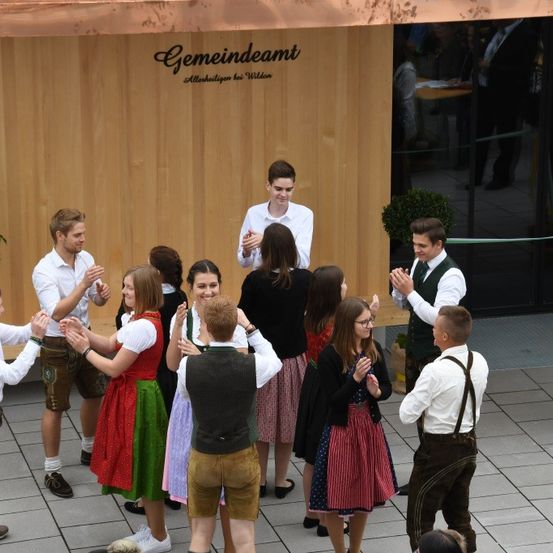 Eine Gruppe von Menschen in traditioneller deutscher Kleidung tanzt vor einem Gebäude namens 'Gemeindeamt'. Einige klatschen, während andere in Gespräche verwickelt sind.