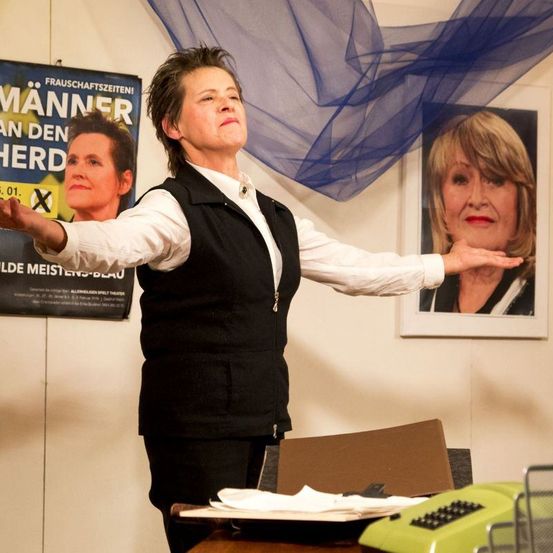 Eine Frau in einer schwarzen Weste steht mit ausgestreckten Armen vor einem Schreibtisch mit einem grünen Telefon. Hinter ihr hat eine Wand einen Poster und ein gerahmtes Bild einer Frau.