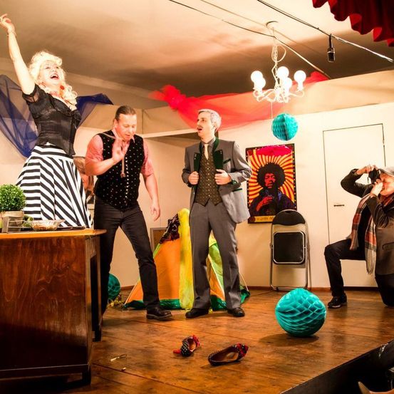 Eine Gruppe von Menschen steht auf der Bühne, eine Person hebt die Hand, während eine andere ein Foto macht. Es gibt einen Schreibtisch mit einer Pflanze darauf und ein Gemälde an der Wand.