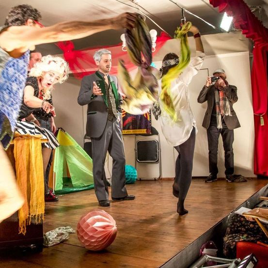 Eine Gruppe von Menschen führt einen theatralischen Akt auf einer hölzernen Bühne auf. Eine Person in einem weißen Hemd schwebt in der Luft. Ein Mann in einem Anzug spricht das Publikum an. Ein Fotograf hält den Moment fest.