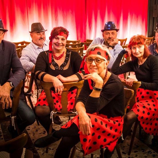 Eine Gruppe von Menschen in roten Punktenröcken und Hüten sitzt auf Stühlen vor einem roten Vorhang.