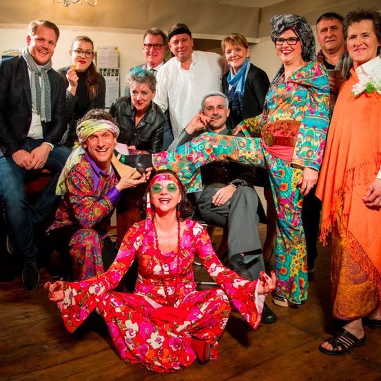 Eine Gruppe von Menschen in farbenfroher Kleidung posiert für ein Foto, wobei eine Frau auf dem Boden sitzt, lächelt und ein Mann ihr Bein hält.