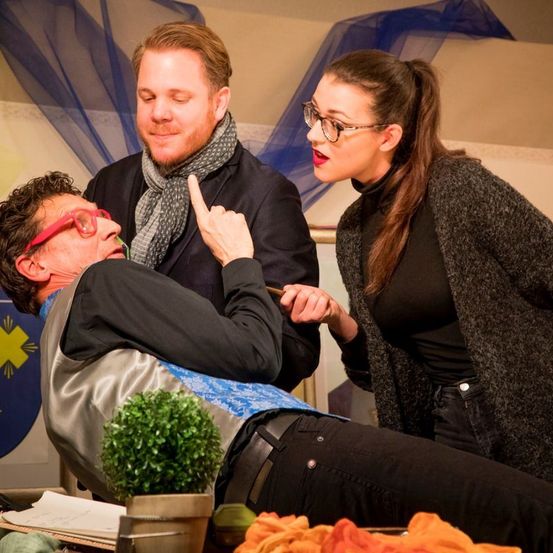 Drei Personen in einem Raum. Ein Mann mit Brille zeigt auf einen anderen Mann, während dieser auf dem Boden liegt. Eine Frau mit Brille sieht sie an.