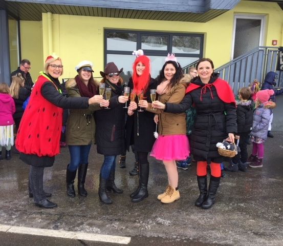 Eine Gruppe von Frauen in Kostümen stoßen mit Champagner vor einem gelben Gebäude mit Treppen und einem Schild an. Kinder im Hintergrund.