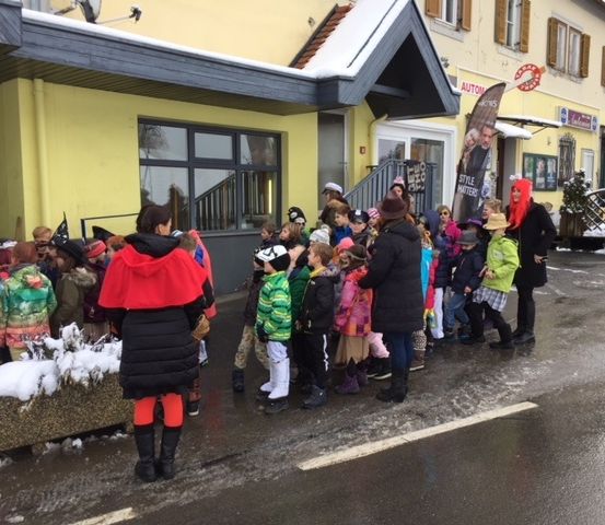 Eine Gruppe von Kindern und Erwachsenen steht auf einer verschneiten Straße, einige tragen Mützen und Stiefel. Vorne eine Frau in einem roten Umhang. Dahinter ein Gebäude mit Glasfenstern und einer Treppe.