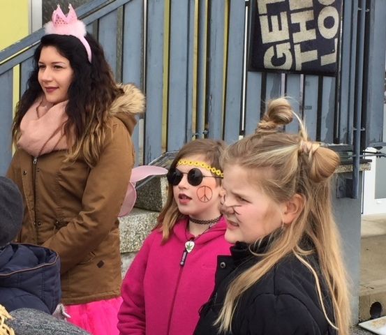 Eine Frau mit einer rosa Krone und einem Schal steht mit zwei Mädchen vor einem blauen Zaun. Ein Mädchen trägt eine Sonnenbrille und ein Friedenszeichen auf der Wange.