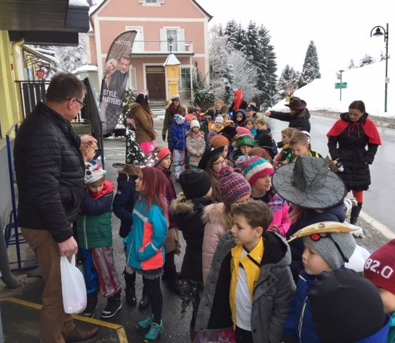 Eine Gruppe von Kindern in Winterkleidung steht vor einem Gebäude, einige tragen Hüte und andere haben Taschen bei sich, während ein Mann einen Beutel vor ihnen hält und ein Banner mit einem Bild eines Mannes dahinter.