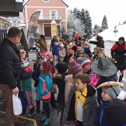 Eine Gruppe von Kindern in Winterkleidung steht vor einem Gebäude, einige tragen Hüte und andere haben Taschen bei sich, während ein Mann einen Beutel vor ihnen hält und ein Banner mit einem Bild eines Mannes dahinter.