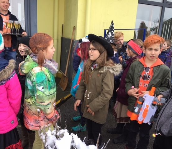 Eine Gruppe von Kindern in Kostümen steht vor einem Gebäude. Einige halten Spielzeugpistolen. Ein Mann mit einem Tablett mit Gläsern steht in der Nähe.