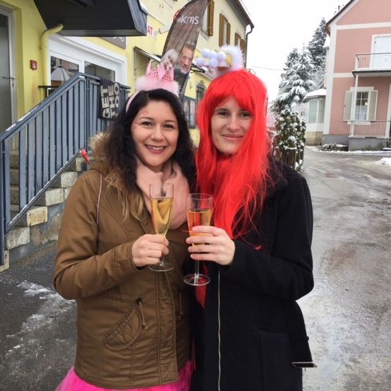 Zwei Frauen in festlicher Kleidung stehen auf einer verschneiten Straße, lächeln und halten Weingläser in den Händen. Hinter ihnen ist ein Gebäude mit einem Banner und einer Treppe zu sehen, und ein Baum mit Schnee ist im Hintergrund.