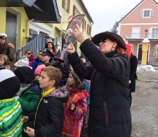 Eine Frau steht vor einer Gruppe von Kindern und hält ein Schild mit einem Bild einer Frau und dem Wort Stil. Die Kinder tragen Winterkleidung und einige haben Mützen auf. Hinter ihnen stehen Gebäude mit Vordächern.